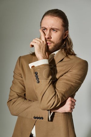 portrait of handsome businessman with long hair and beard posing in beige blazer on grey backdrop