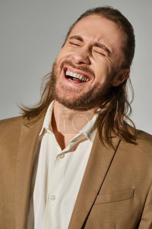 portrait of handsome businessman with long hair and beard laughing with closed eyes on grey backdrop