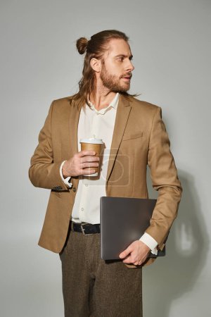 Photo for Handsome and bearded businessman in formal attire holding laptop and coffee on grey backdrop - Royalty Free Image