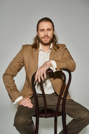 handsome bearded businessman in beige formal attire sitting on wooden chair on grey background