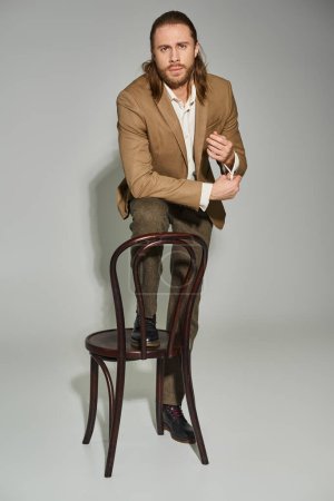 attractive bearded businessman in beige formal attire posing near wooden chair on grey background