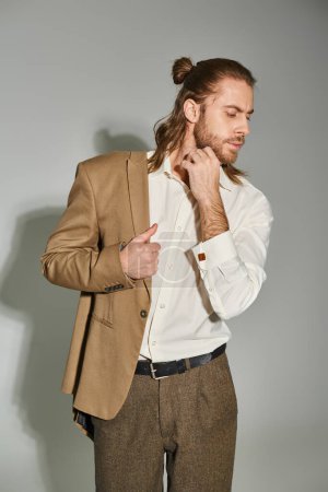 hombre de negocios barbudo bien parecido en camisa blanca posando con chaqueta beige en el hombro en gris