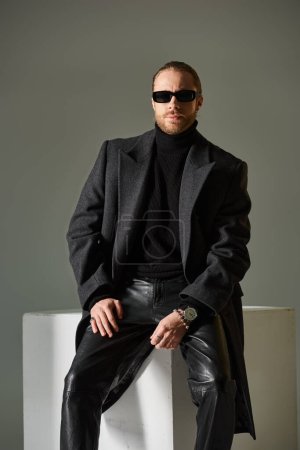 handsome bearded man in stylish sunglasses and woolen coat posing near white cube on grey backdrop