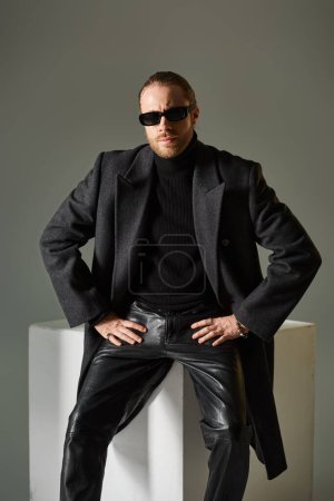gutaussehender bärtiger Mann mit stylischer Sonnenbrille und Mantel sitzt auf weißem Würfel vor grauem Hintergrund