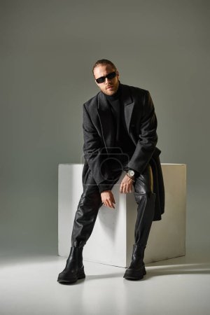 full length of bearded man in stylish sunglasses and coat sitting on white cube on grey backdrop
