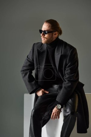 good looking stylish man in trendy sunglasses and coat sitting on white cube on grey backdrop
