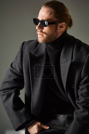 portrait of good looking stylish man in sunglasses and coat sitting on white cube on grey backdrop