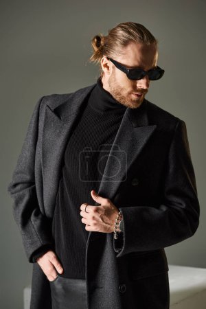 Porträt eines modischen Mannes mit trendiger Sonnenbrille und Wollmantel, der auf grauem Hintergrund posiert
