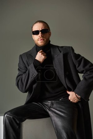 portrait of fashionable model in trendy sunglasses and coat sitting on white cube on grey backdrop