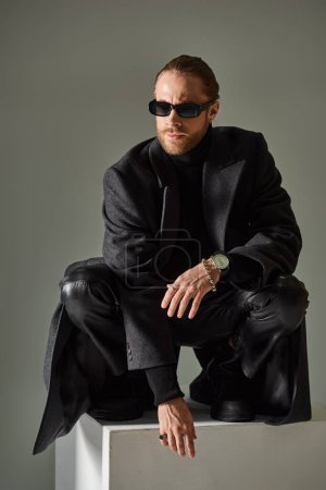 fashionable male model in trendy sunglasses and coat sitting on top of on white cube on grey