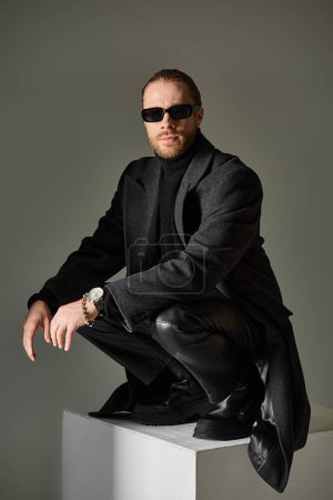 fashionable male model in trendy sunglasses and black attire sitting on top of on white cube on grey