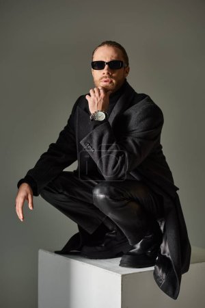 stylish male model in trendy sunglasses and black attire sitting on top of on white cube on grey
