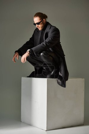 handsome male model in trendy sunglasses and black attire sitting on top of on white cube on grey