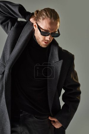 portrait of handsome male model in trendy sunglasses and black attire standing on grey backdrop