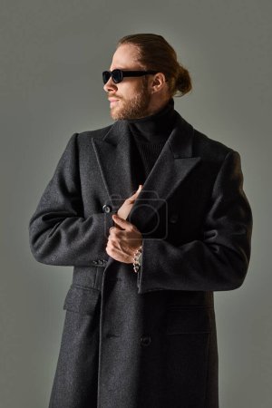 Porträt eines hübschen männlichen Modells mit trendiger Sonnenbrille und schwarzer Kleidung, das vor grauem Hintergrund wegschaut
