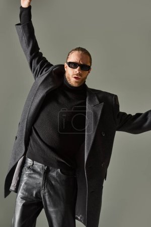 Porträt eines gutaussehenden bärtigen Mannes mit trendiger Sonnenbrille und schwarzer Kleidung, der mit erhobenen Händen posiert