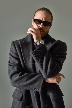 Foto de Hombre elegante y barbudo en gafas de sol oscuras de moda y traje negro posando sobre fondo gris - Imagen libre de derechos
