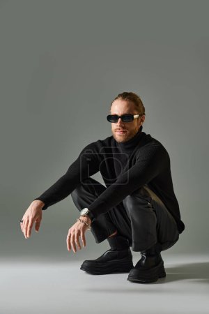 Photo for Bearded man in trendy sunglasses, turtleneck sweater and leather pants sitting on haunches on grey - Royalty Free Image