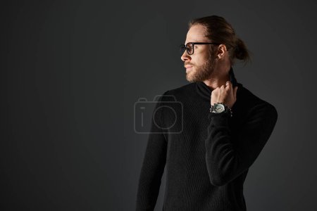 handsome man in eyeglasses pulling high collar on turtleneck sweater while posing on grey background