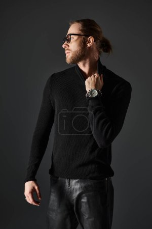bearded man in eyeglasses pulling high collar on turtleneck sweater while posing on grey background