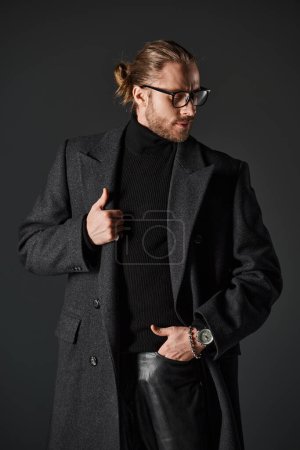 bearded man in eyeglasses and classy woolen coat posing with hand in pocket on grey background