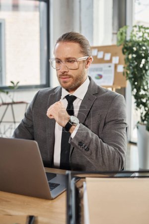 Nachdenklicher Geschäftsmann in grauem Anzug und Brille arbeitet in modernen Büroräumen am Laptop