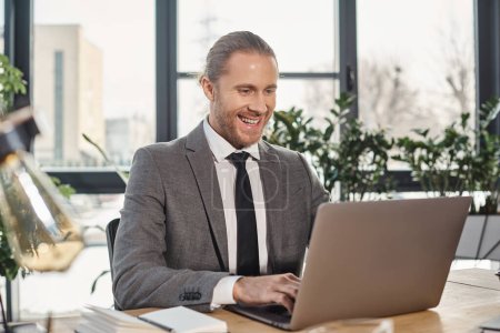 fröhlicher und stilvoller Geschäftsmann im grauen Anzug tippt auf Laptop während er im modernen Büro arbeitet