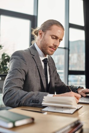 focused businessman in grey suit writing in notebook near laptop in modern work environment