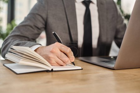 vista recortada del hombre de negocios en la escritura de ropa formal en el cuaderno mientras está sentado en el lugar de trabajo en la oficina