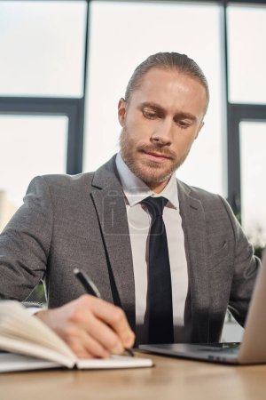 homme d'affaires sérieux et ambitieux en gris assis près de l'ordinateur portable et l'écriture dans le carnet, la productivité