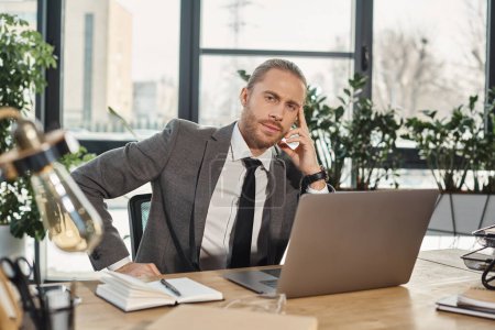 serious stylish businessman sitting at workplace near laptop and looking at camera in office