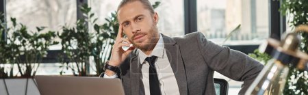 empresario pensativo en traje gris sentado en el lugar de trabajo cerca de la computadora portátil y mirando a la cámara, pancarta