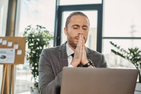 Photo for Worried businessman sitting with praying hands and looking at laptop while working in office - Royalty Free Image