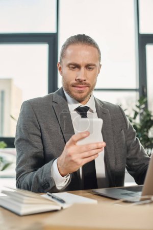Nachdenklicher Geschäftsmann im grauen Anzug sitzt neben Laptop auf Schreibtisch und schaut aufs Smartphone