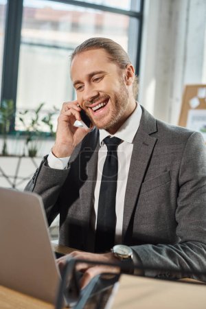 fröhlicher stilvoller Geschäftsmann, der am Arbeitsplatz in modernem Büro am Smartphone neben dem Laptop spricht