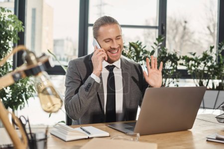 joyful businessman talking on smartphone and waving hand during video chat on laptop in office