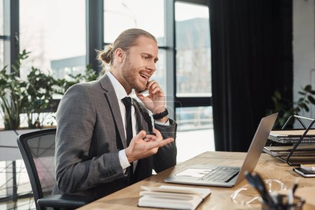 nervöser Geschäftsmann zeigt auf Laptop, während er am Arbeitsplatz im Büro mit dem Handy telefoniert