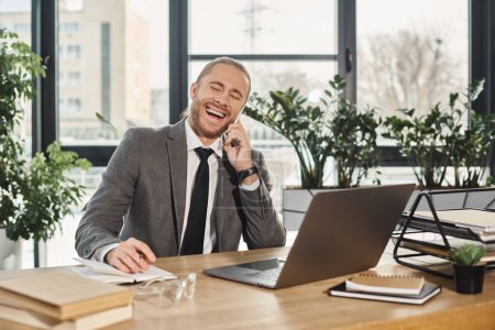 emocionado empresario exitoso hablando en el teléfono inteligente y riendo cerca de la computadora portátil en la oficina moderna
