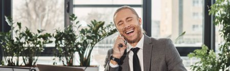 regocijado exitoso hombre de negocios riendo durante la conversación en el teléfono inteligente en la oficina, bandera