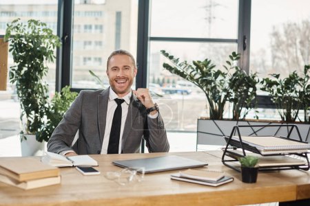 Photo for Cheerful businessman looking at camera at work desk with documents and digital devices in office - Royalty Free Image
