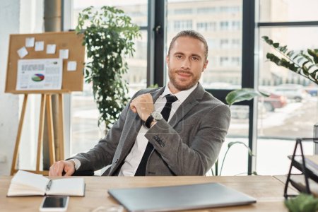 Ambitionierter Geschäftsmann im grauen Anzug blickt im Büro in die Kamera neben Laptop und Smartphone