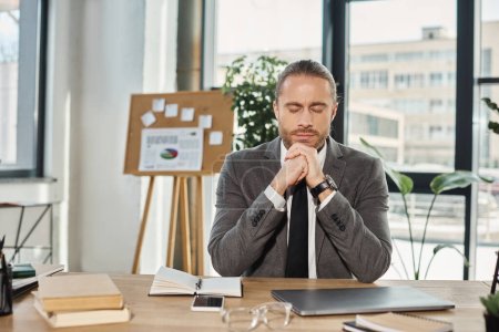 Müder Geschäftsmann sitzt mit geschlossenen Augen neben Laptop und Smartphone am Schreibtisch im modernen Büro