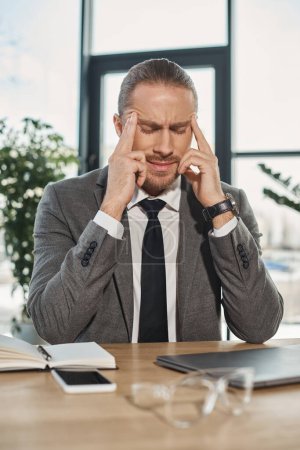 Müder Geschäftsmann, der unter Kopfschmerzen leidet und mit geschlossenen Augen am Arbeitsplatz im modernen Büro sitzt