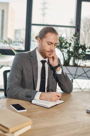Photo for Thoughtful businessman in formal wear writing in notebook near smartphone on work desk in office - Royalty Free Image