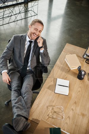 top view of happy businessman talking on smartphone and smiling with closed eyes at workplace