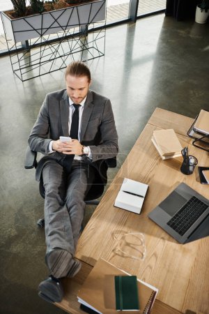 top view of manager sitting in relaxed pose and messaging on smartphone near notebooks and laptop