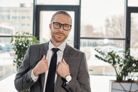 Porträt eines positiven Geschäftsmannes in grauem Anzug und Brille, der im modernen Büro in die Kamera lächelt