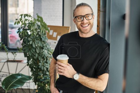 cheerful businessman in black t-shirt and eyeglasses holding coffee to go and looking away in office