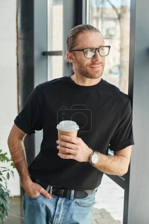 gerente sonriente en camiseta negra y anteojos sosteniendo bebida para llevar y mirando hacia otro lado en la oficina