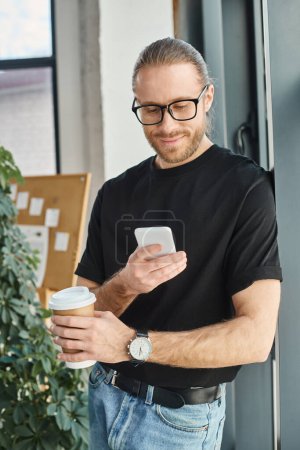 happy businessman in eyeglasses and casual clothes looking at smartphone and holding coffee to go